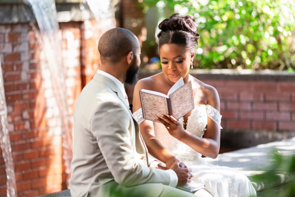 Seated together near a brick fountain, the bride holds a floral-patterned notebook and reads aloud while the groom listens attentively, their hands clasped. This private vow exchange takes place in the quiet garden space at the Waterhouse Pavilion, surrounded by soft greenery and flowing water.