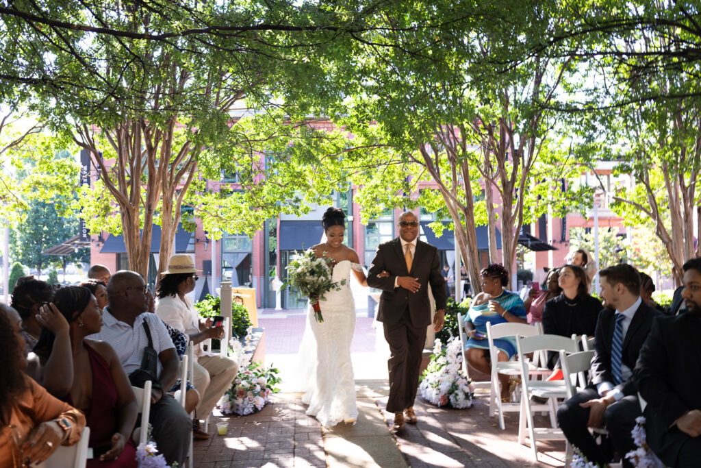 The bride walks arm in arm with her father down a flower-lined aisle, framed by an archway of leafy trees. Guests turn in their chairs to watch as she approaches the altar, with the open-air beauty of the Waterhouse Pavilion creating an elegant backdrop to her entrance.