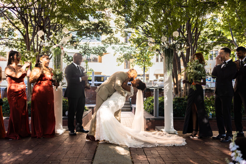 Cheers erupt from the wedding party as the groom dips the bride into a dramatic kiss, framed by tall floral columns and leafy trees. The lively celebration reflects the joy of a downtown Chattanooga wedding venue, where city charm meets natural beauty.