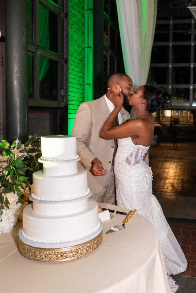 The couple leans into a joyful kiss beside their elegant three-tier white wedding cake, decorated simply with a gold-trimmed base and surrounded by greenery. The glow of evening uplighting inside the Waterhouse Pavilion casts a warm ambiance over this sweet cake-cutting moment.