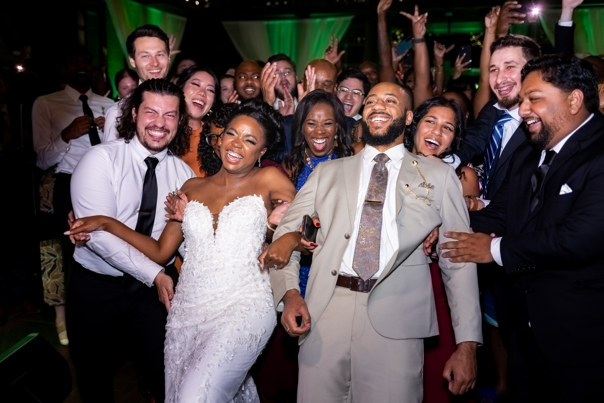 Surrounded by friends, the newlyweds laugh and lean back into a crowd of cheering guests on the dance floor. The group’s energy fills the reception space, with the Waterhouse Pavilion alive in celebration, colorful lights reflecting off smiling faces.