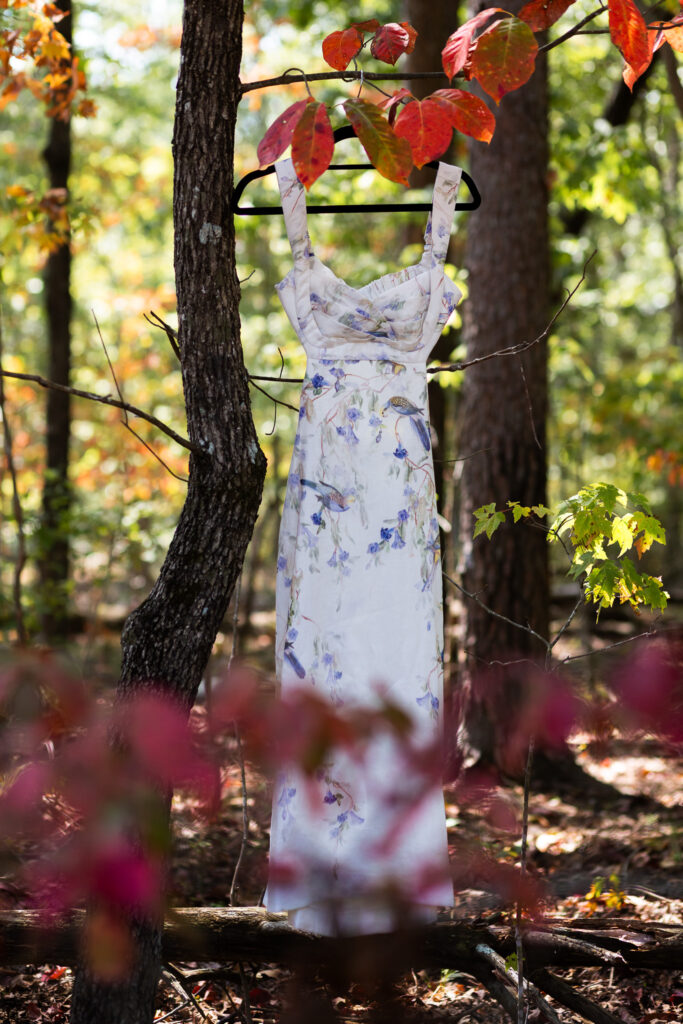 A floral wedding dress with soft purple and green details hangs from a tree branch surrounded by autumn leaves, setting the scene for a Georgia State Park wedding in the middle of nature.