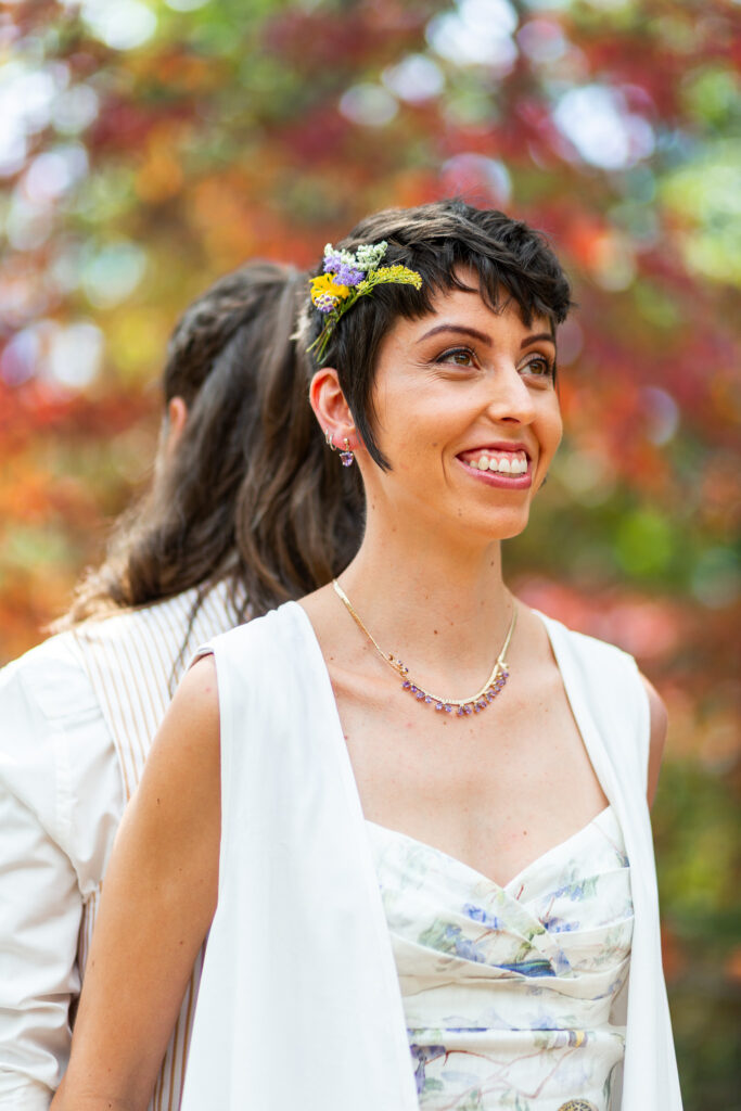 The bride smiles softly, wearing a floral crown and delicate necklace, as the autumn leaves blur into the background of this Georgia State Park wedding first look.