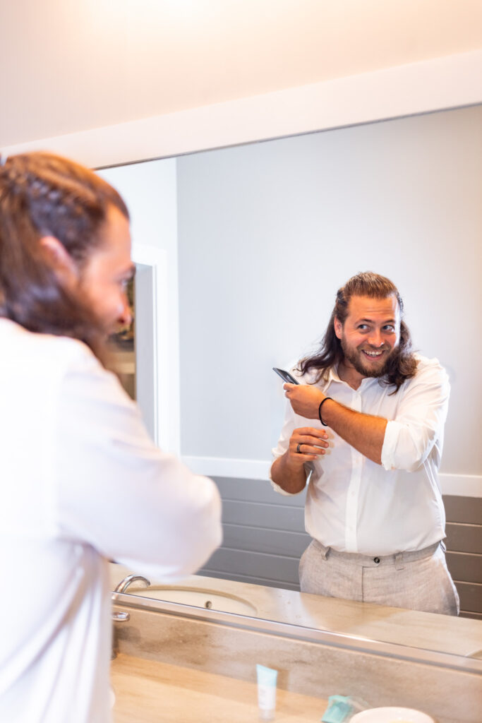 The groom smiles at his reflection in the mirror while brushing his hair in a bright, modern cabin bathroom before the Georgia State Park wedding begins.