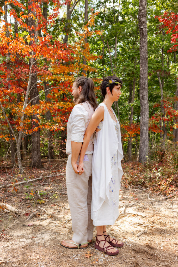 The couple stands back-to-back in the forest, holding hands before seeing one another, surrounded by vibrant fall colors at their Georgia State Park wedding.