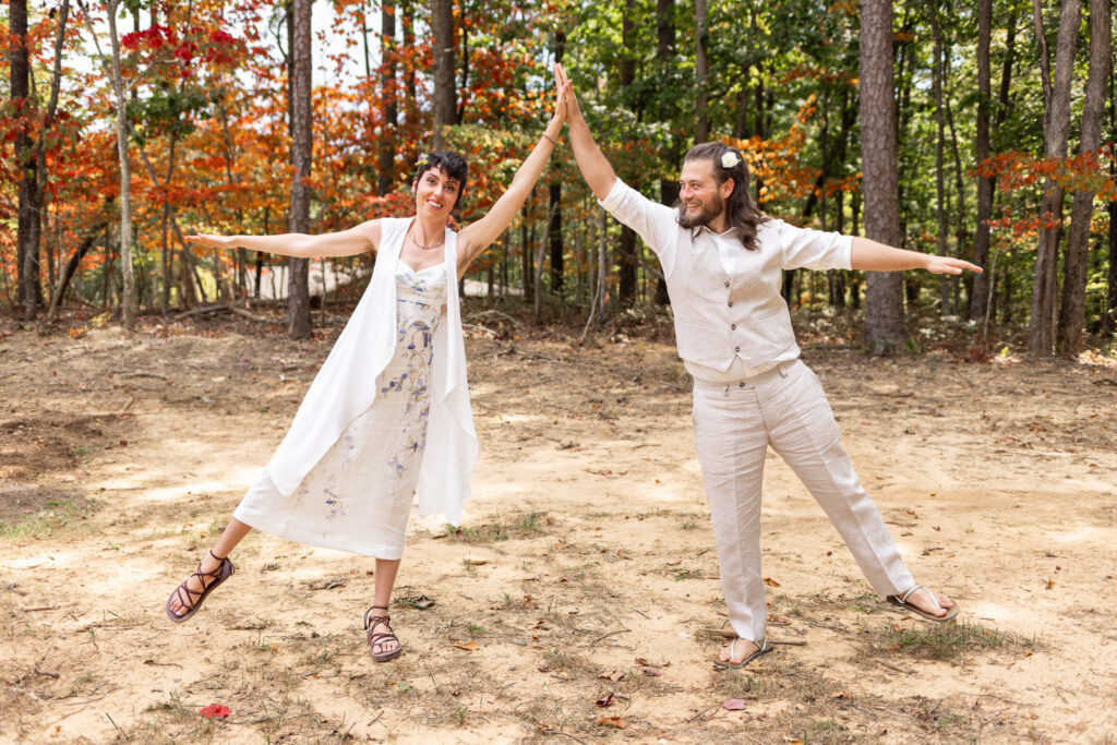 Two figures balance like airplanes in the woods, leaning outward with arms extended as their hands meet in a high five. Their linen outfits sway slightly, and the orange leaves scattered among the trees create a frame of warmth around their joy-filled pose.