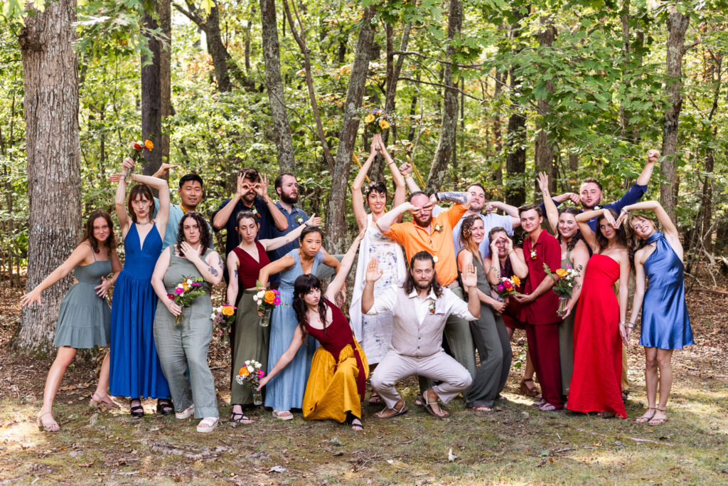 The entire wedding party gathers in the woods, striking playful and exaggerated poses. Colorful dresses and suits stand out against the green trees, bouquets held aloft as laughter fills the frame in this joyful group portrait.