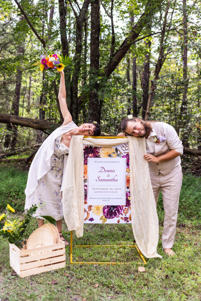 A bright welcome sign framed with soft draped fabric stands in a clearing, surrounded by tall trees and a wooden crate of sunflowers at its base. The couple leans playfully against it, one lifting a bouquet high in the air. The carefree energy of this moment reflects the colorful spirit of a Cloudland Canyon wedding set deep in the forest.