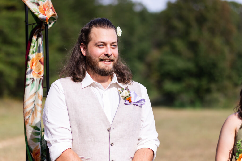 The groom stands at the altar, eyes wide and soft with emotion as he looks forward. Sunlight highlights the flower tucked into his hair and the textured linen of his vest, while a fabric-draped arch frames the moment just behind him.