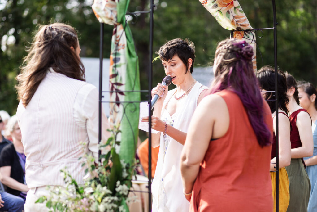 The bride reads vows into a microphone, her eyes on the small sheet of paper in her hand. The fabric arch sways gently behind her, while attendants listen with quiet focus. Surrounded by forest and loved ones, the words carry through the air, embodying the intimacy of a Cloudland Canyon wedding.
