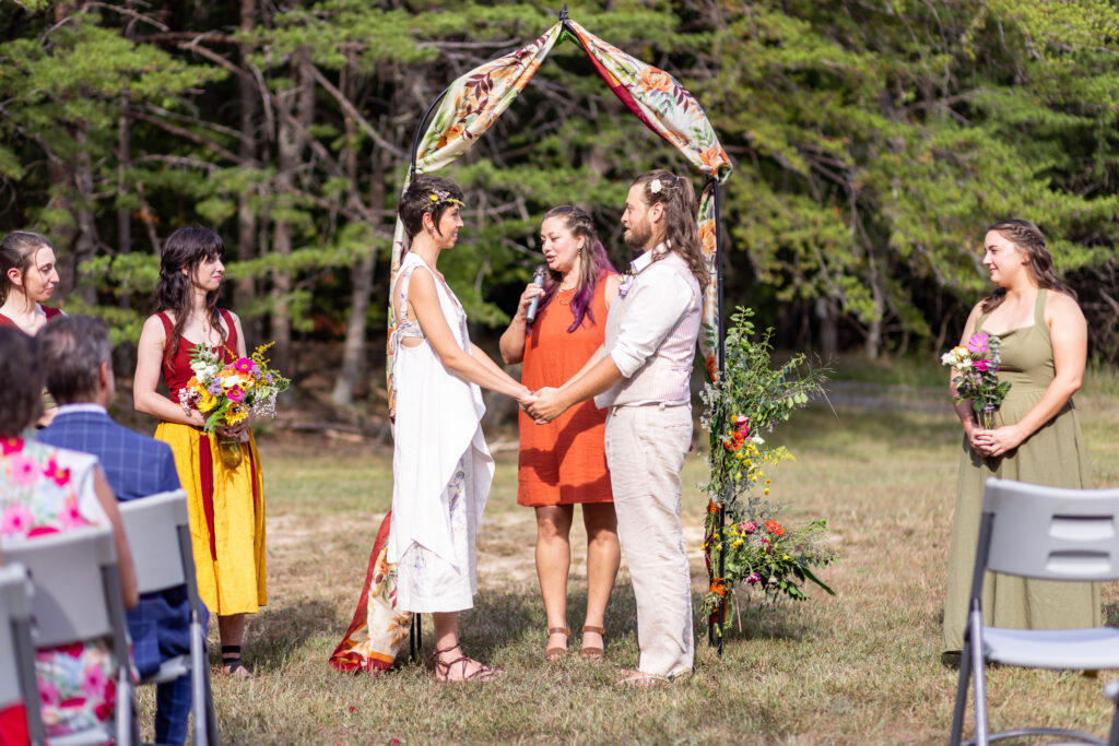 The couple stands hand in hand beneath a vibrant arch of wildflowers and fabric, their gazes locked as the officiant speaks. Attendants holding mason jar bouquets stand nearby, adding to the layered, colorful details that make this Cloudland Canyon wedding both grounded and full of life.