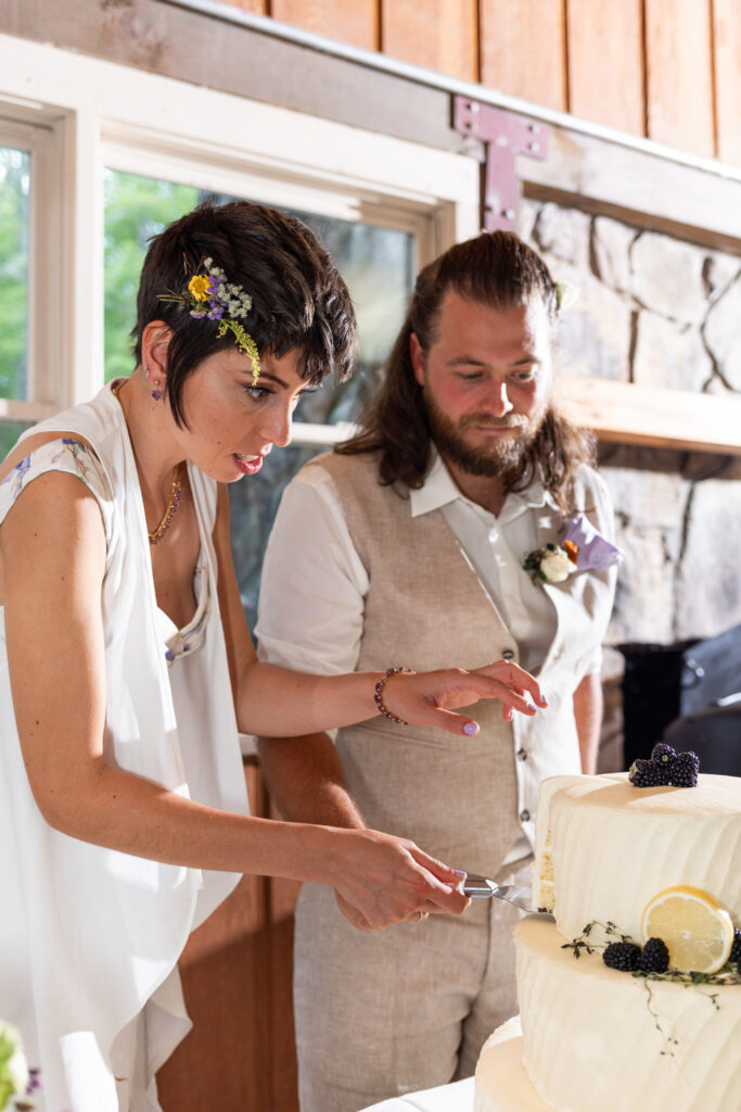 The glow of daylight streams through cabin windows as the couple cuts into a three-tiered cake decorated with lemon slices and blackberries. Focused expressions fill their faces as one guides the knife carefully into the bottom layer, the texture of the frosting catching the light.