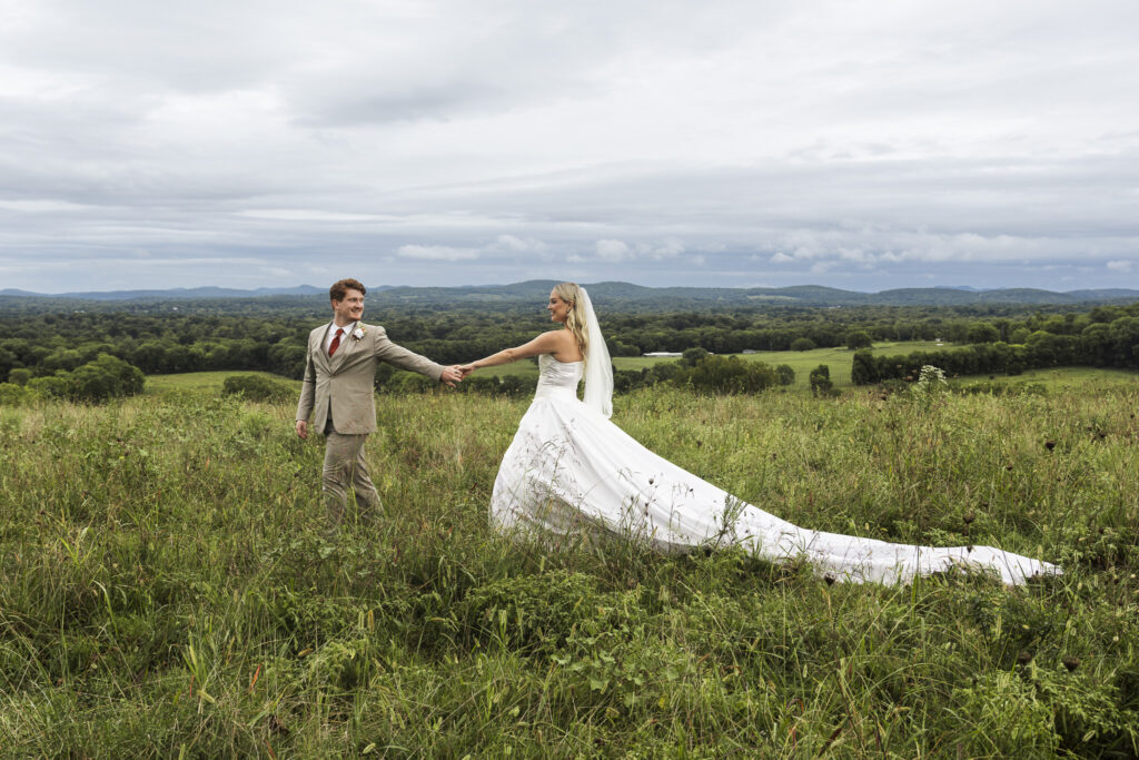 Bride and groom walking hand in hand through a scenic field— Her extra long train flowing behind her, a testament to her wedding dress planning and portrait session.