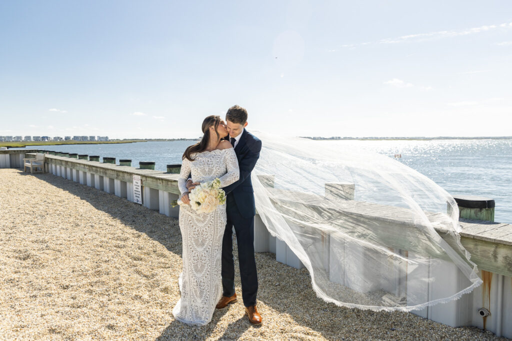 Bride and groom embracing by the water with veil blowing in the wind, showing the beauty and movement achieved through wedding dress planning.
