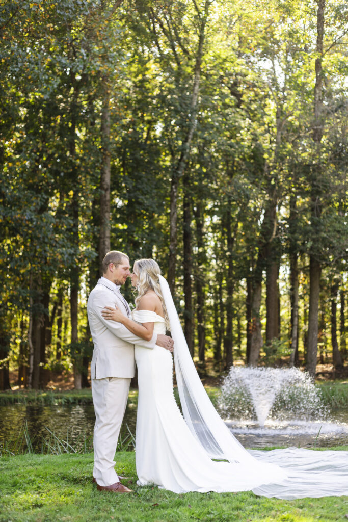 Standing by a tranquil pond and fountain, the couple shares a quiet moment together, sunlight filtering through the trees during their romantic Lehigh Valley wedding portraits.