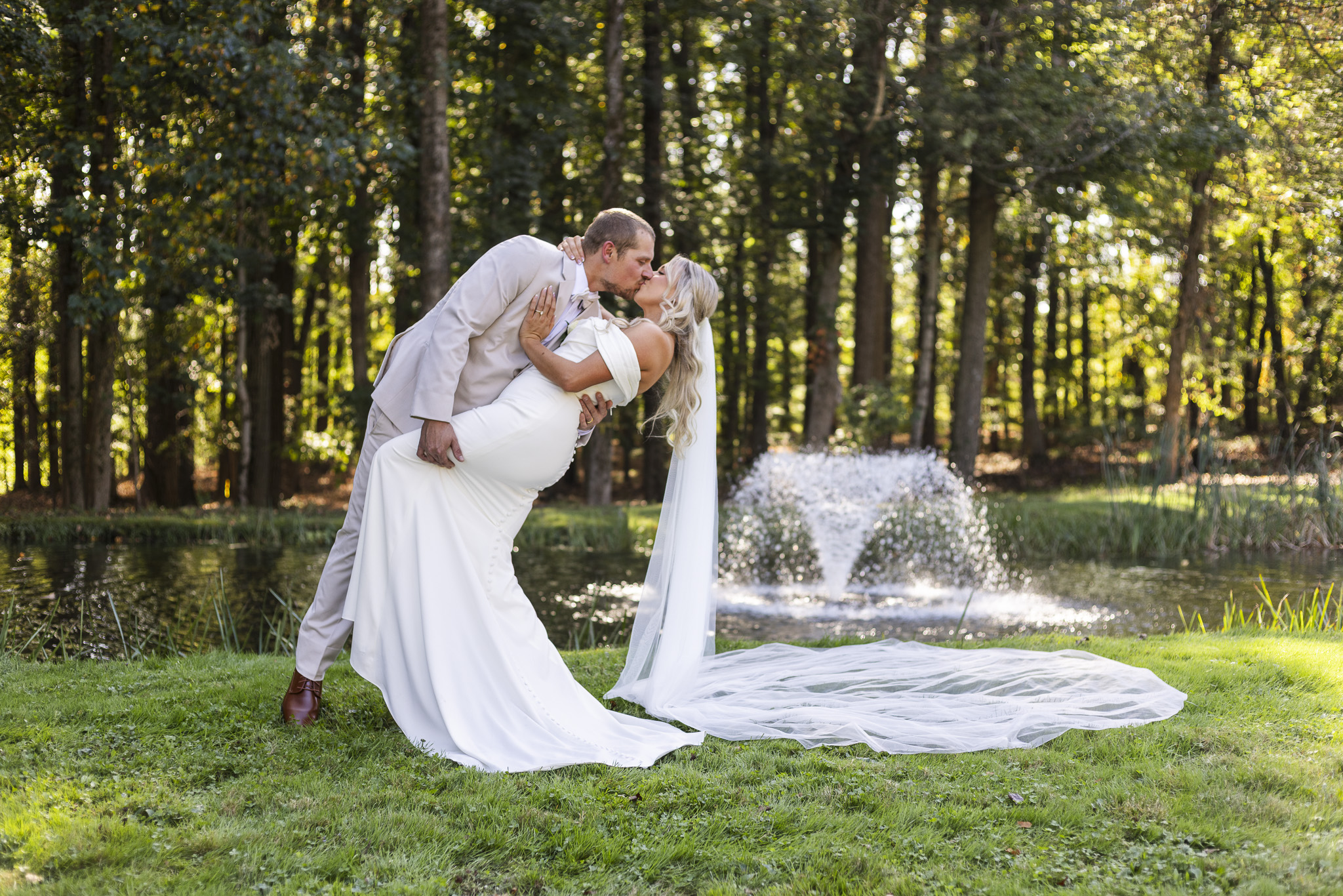 The groom dips the bride into a romantic kiss beside a pond, her long veil flowing across the grass as the fountain sprays softly behind them in the late afternoon light.