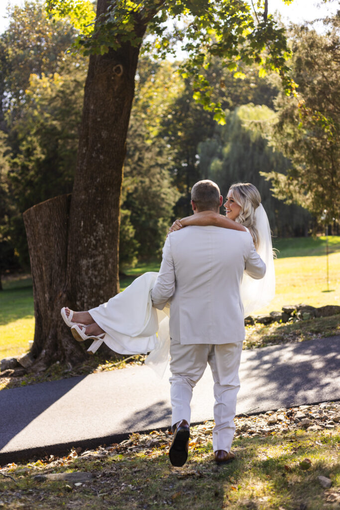 The groom carries his bride through the gardens, her dress trailing softly in the sunlight as they share a quiet walk together after their Domaine Pterion wedding ceremony.