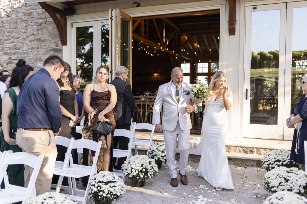 The bride walks arm-in-arm with her father, smiling through tears as she makes her way down the aisle surrounded by guests at this Domaine Pterion wedding celebration.