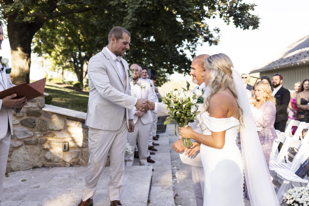 As the bride’s father gives her away, the groom reaches out to shake his hand, marking a touching transition during this Domaine Pterion wedding ceremony.