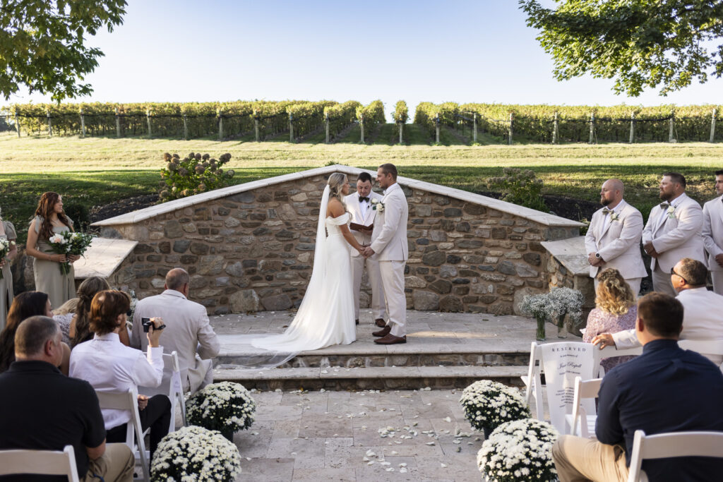 The couple holds hands before a stone wall and vineyard backdrop, exchanging vows under the open sky during this Domaine Pterion wedding in the Lehigh Valley.