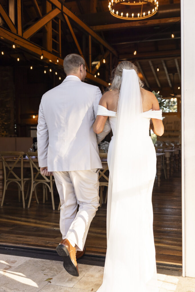 Arm in arm, the newlyweds step into the warm glow of the reception barn, soft string lights illuminating the wooden beams overhead.