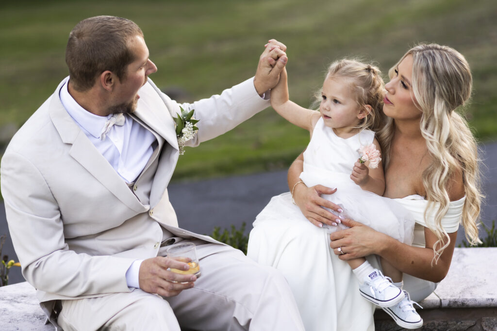 The bride sits with a small flower girl on her lap while the groom playfully raises her hand for a high five, sharing a relaxed and joyful family moment after the ceremony.