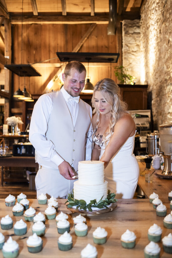 Inside the rustic stone barn, the newlyweds cut their tiered white cake surrounded by cupcakes, laughing together during their Domaine Pterion wedding reception.