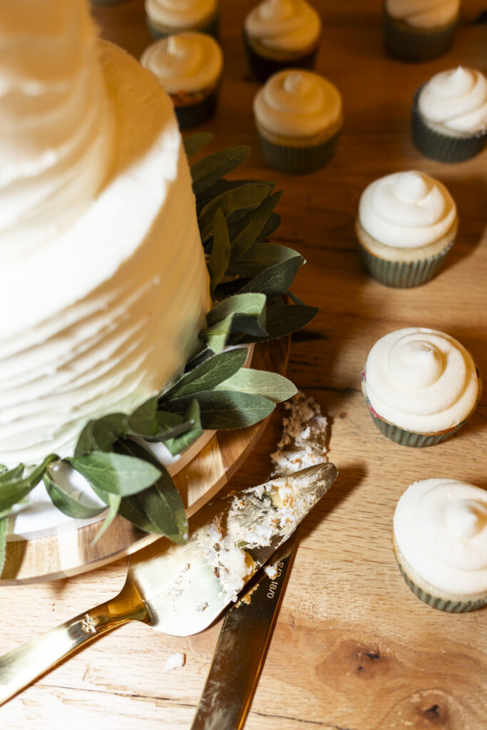 A close-up of the dessert table shows crumbs and frosting left on a gold cake knife after the couple’s cake cutting, surrounded by vanilla cupcakes topped with buttercream.