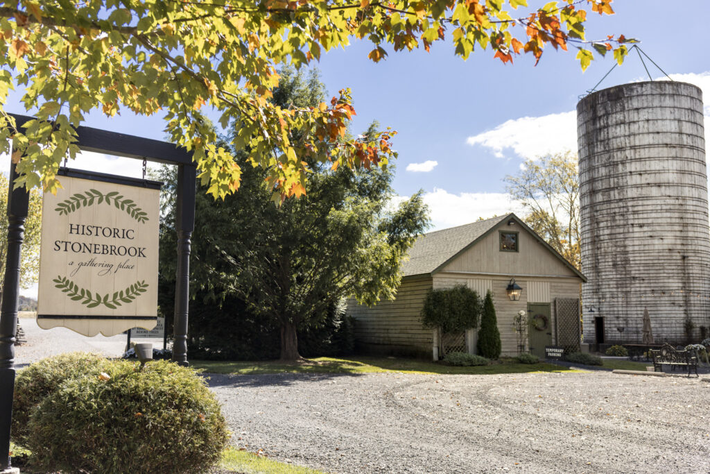 A view of Historic Stonebrook Farm’s iconic white silo and weathered barn framed by autumn leaves under a bright blue sky, setting the rustic scene for a charming Historic Stonebrook wedding celebration.