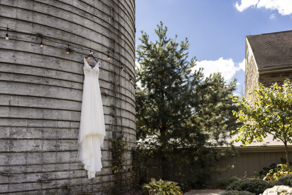 A flowing chiffon and lace wedding gown hangs gracefully from the curved wall of a weathered silo at a Bucks County wedding venue, illuminated by string lights and framed by evergreens and blue skies at Historic Stonebrook Farm.