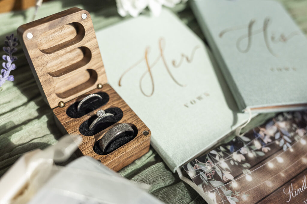 A wooden ring box filled with engraved silver bands and a round-cut diamond engagement ring sits beside sage green vow books and lavender sprigs, styled beautifully for a rustic Bucks County wedding at Historic Stonebrook Farm.