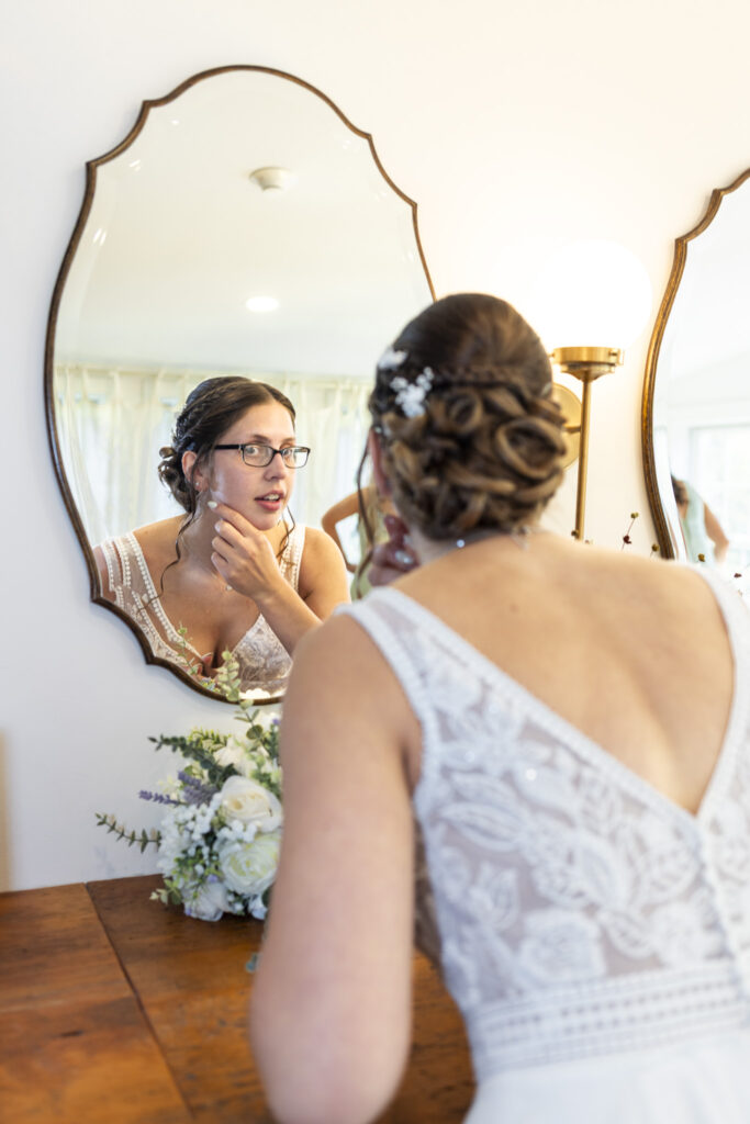 Captured in front of a vintage mirror, the bride applies her final touches with calm focus, her intricate updo adorned with small white flowers and her bouquet resting nearby—an intimate glimpse from a Bucks County wedding morning.
