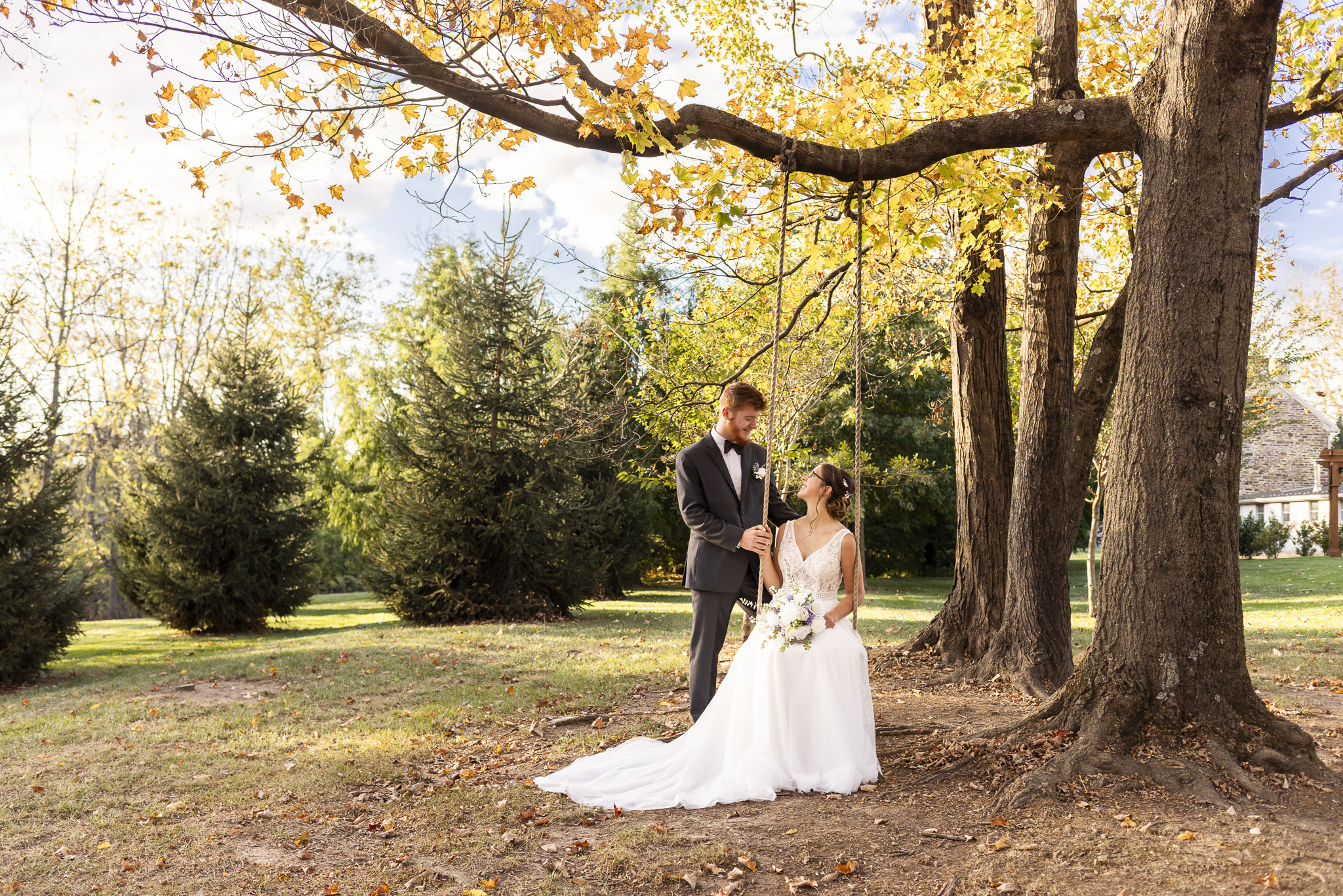 Beneath golden autumn leaves, the groom stands beside the bride as she sits gracefully on a tree swing, both smiling up at each other surrounded by tall evergreens and late-fall color.