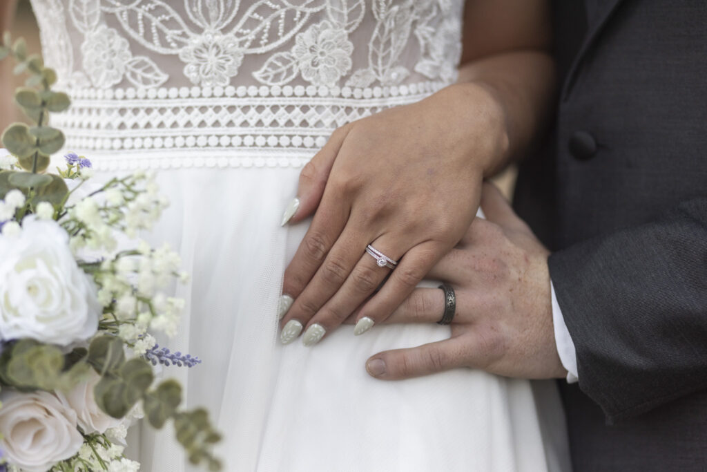 Close-up of the couple’s hands resting over her flowing skirt, the bride’s diamond ring and manicured nails softly highlighted beside the groom’s engraved wedding band and her bouquet of pale lavender and ivory blooms.