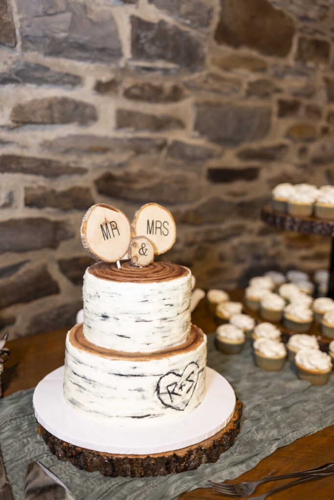 A close-up of the couple’s two-tier birch-inspired cake topped with wooden “Mr. & Mrs.” rounds and surrounded by cupcakes captures the woodland theme of their Historic Stonebrook Farm wedding reception.