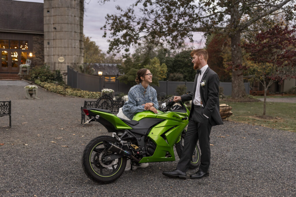 Wearing her lace gown and a pearl-studded denim jacket, the bride laughs beside the groom as she's flirty, leaning on the motorcycle, framed by autumn trees and soft light spilling from the barn during their Historic Stonebrook Farm wedding send-off.