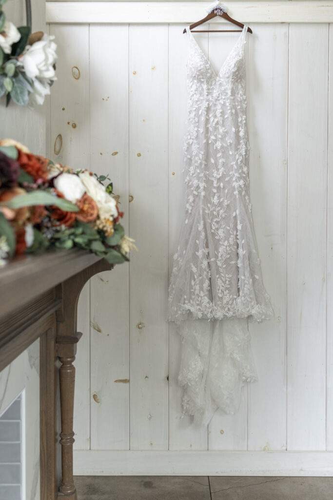 The bride’s lace wedding gown hangs elegantly on a wooden hanger against a white paneled wall, with her bouquet of rust, ivory, and sage florals blurred in the foreground at this Lancaster farmhouse wedding.