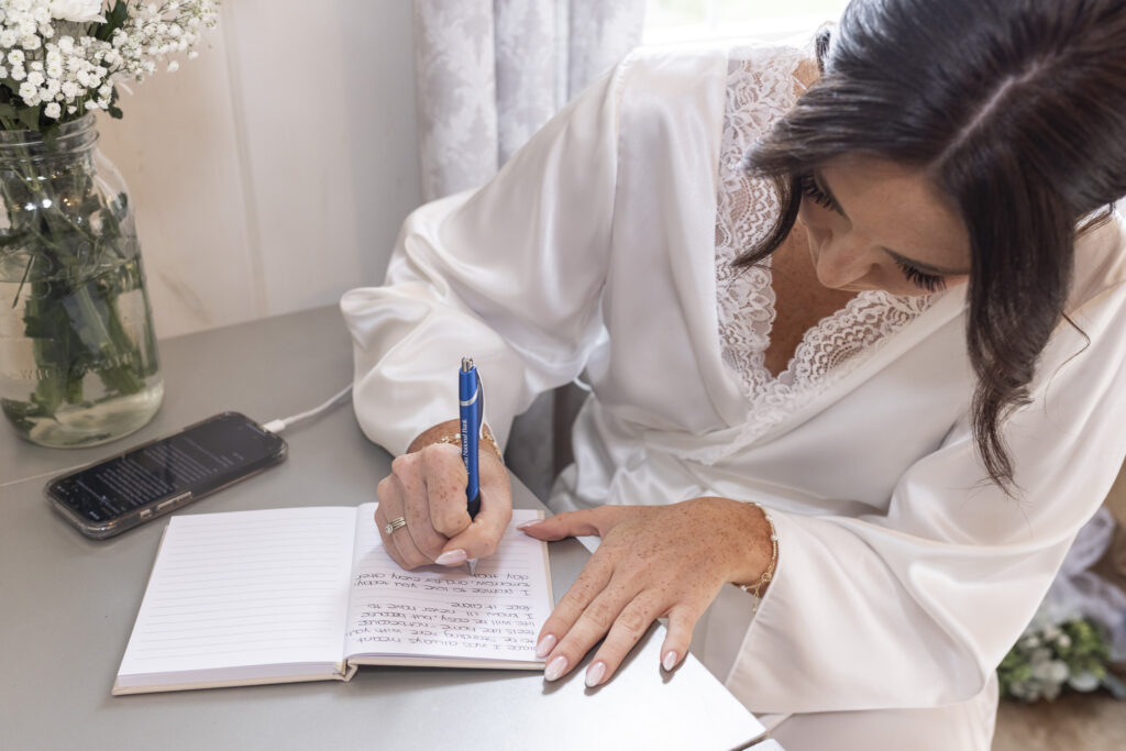 Wearing a white satin robe, the bride leans over her vow book, pen in hand, as she writes heartfelt words before her Lancaster farmhouse wedding ceremony begins.