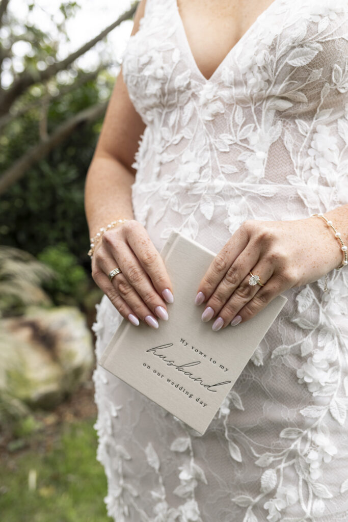 A close-up of the bride’s torso shows her hands gently holding a cream-colored vow book embossed with the words “My vows to my husband on our wedding day.” Her nails are pale pink, and her lace gown is covered in delicate floral appliqués. Soft greenery fills the blurred background.