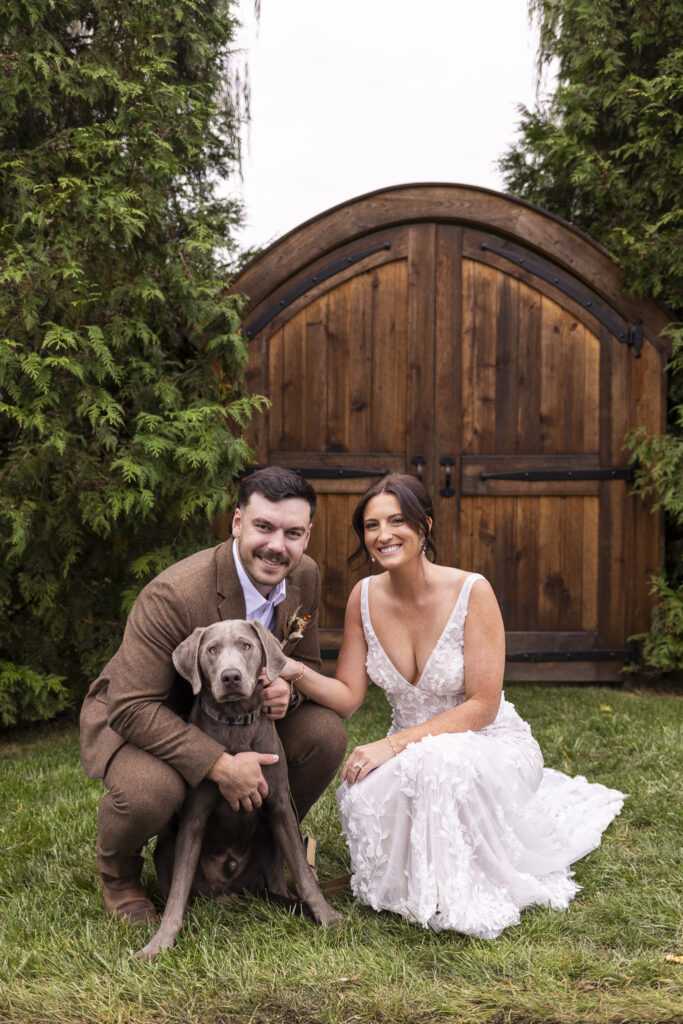 In front of the arched wooden doors framed by evergreens, the bride and groom kneel beside their gray Weimaraner, smiling together during their Stoltzfus Homestead Wedding portraits.
