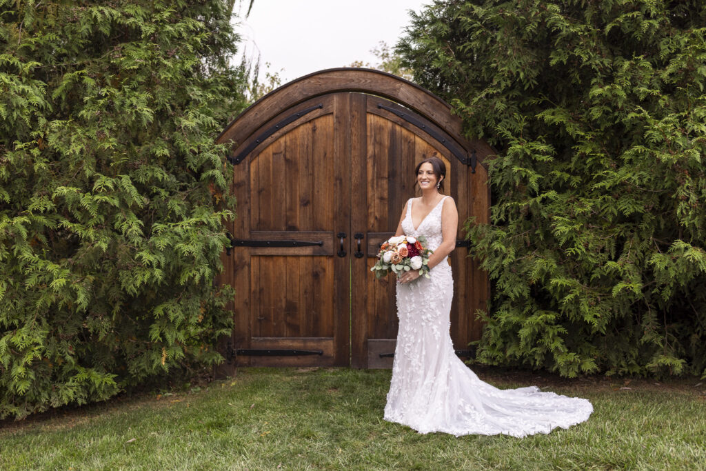 The bride smiles softly as she poses with her bouquet of fall-toned blooms in front of arched wooden doors framed by lush greenery—an iconic backdrop for a Lancaster farmhouse wedding portrait.