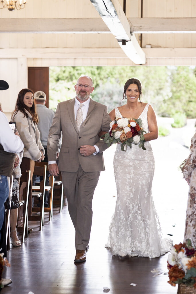 The bride walks arm-in-arm with her father down the aisle of The Stoltzfus Homestead barn, sunlight filtering through open doors as guests rise to greet her with smiles and tears.