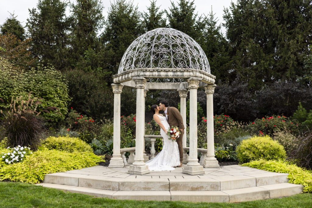 Under a white stone gazebo surrounded by manicured gardens, the couple kisses beneath an overcast sky, their Lancaster farmhouse wedding portraits blending elegance with nature’s warmth.
