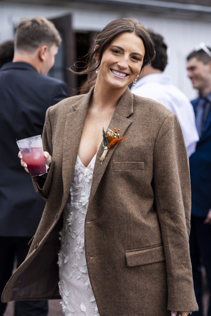 Wrapped in her groom’s tweed jacket, the bride laughs with a drink in hand as guests mingle in the courtyard during a Stoltzfus Homestead wedding cocktail hour, where farmhouse charm meets relaxed autumn celebration.