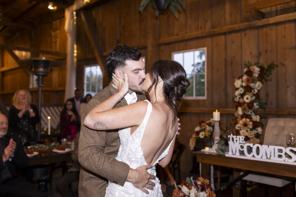 The bride and groom share a slow kiss during their first dance. Her hand cradles the back of his neck while his arms wrap around her waist. Guests watch from wooden tables in the softly lit barn, decorated with candles and fall-colored flowers.