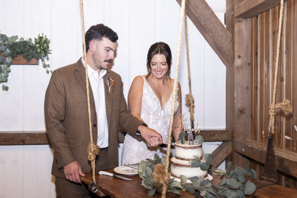 In a cozy barn reception space, the newlyweds cut their semi-naked cake decorated with eucalyptus leaves and wooden accents—an inviting detail from their Lancaster farmhouse wedding celebration.