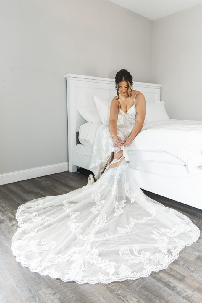 Bride sitting on the edge of a white bed, fastening her wedding shoes while her lace gown flows elegantly across the floor.