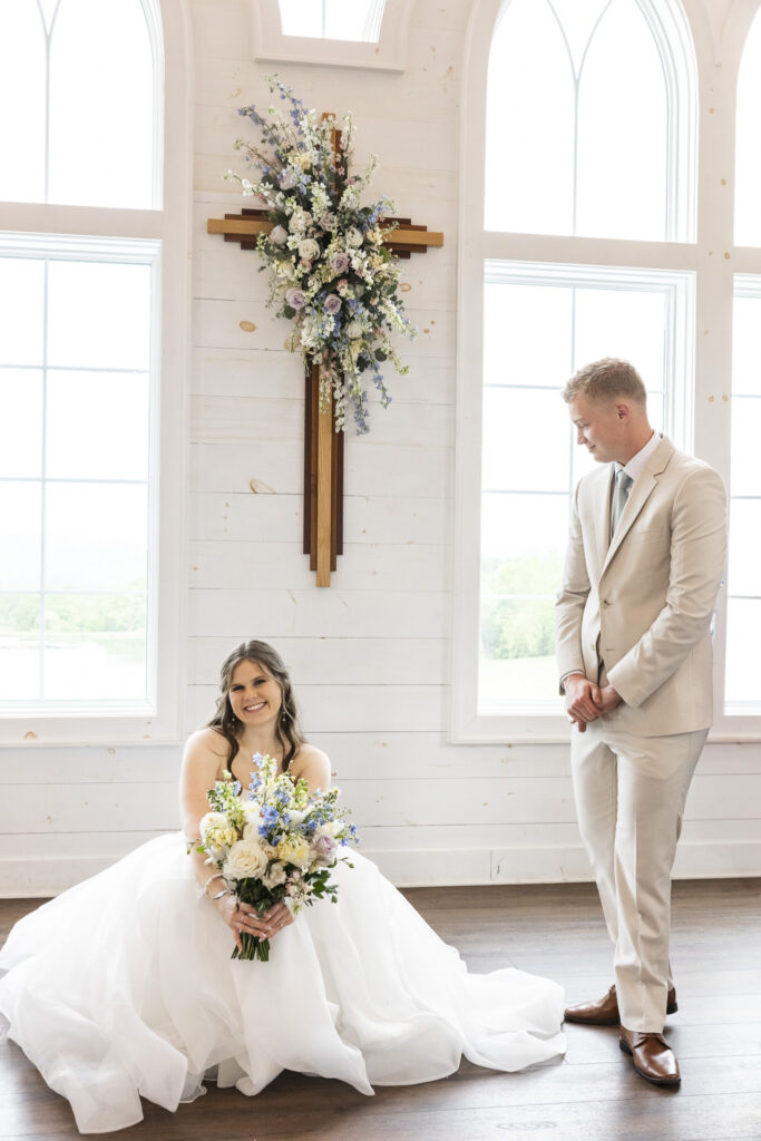 Voluminous tulle wedding gown with layered skirt and sweetheart neckline worn by the bride as she squats next to groom in-between portraits in a white chapel