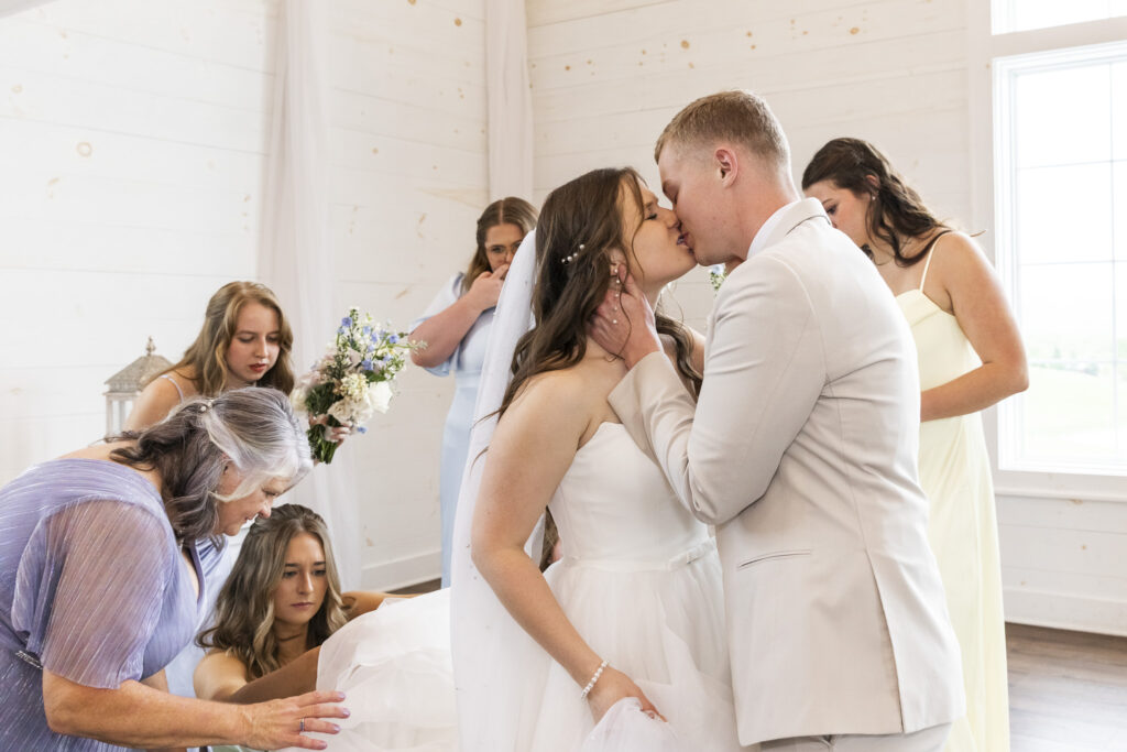 Bride and groom share a kiss as bridesmaids and family help bustle the gown during a joyful wedding moment in the chapel.