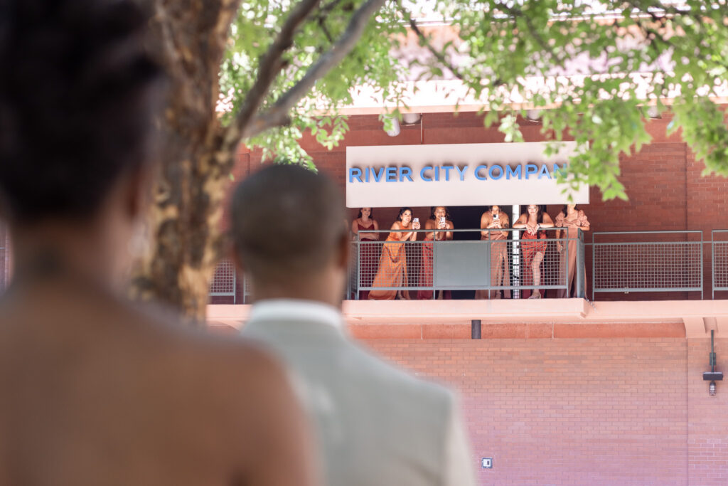 Just before their first look the bride and groom stand in the foreground, their backs slightly blurred as they look up toward the balcony. Above them, the bridesmaids lean over the railing of the River City Company building, holding phones, wiping away tears, and cheering with joy. The moment captures the heartfelt perspective of the first look, framed by trees and warm brick architecture.